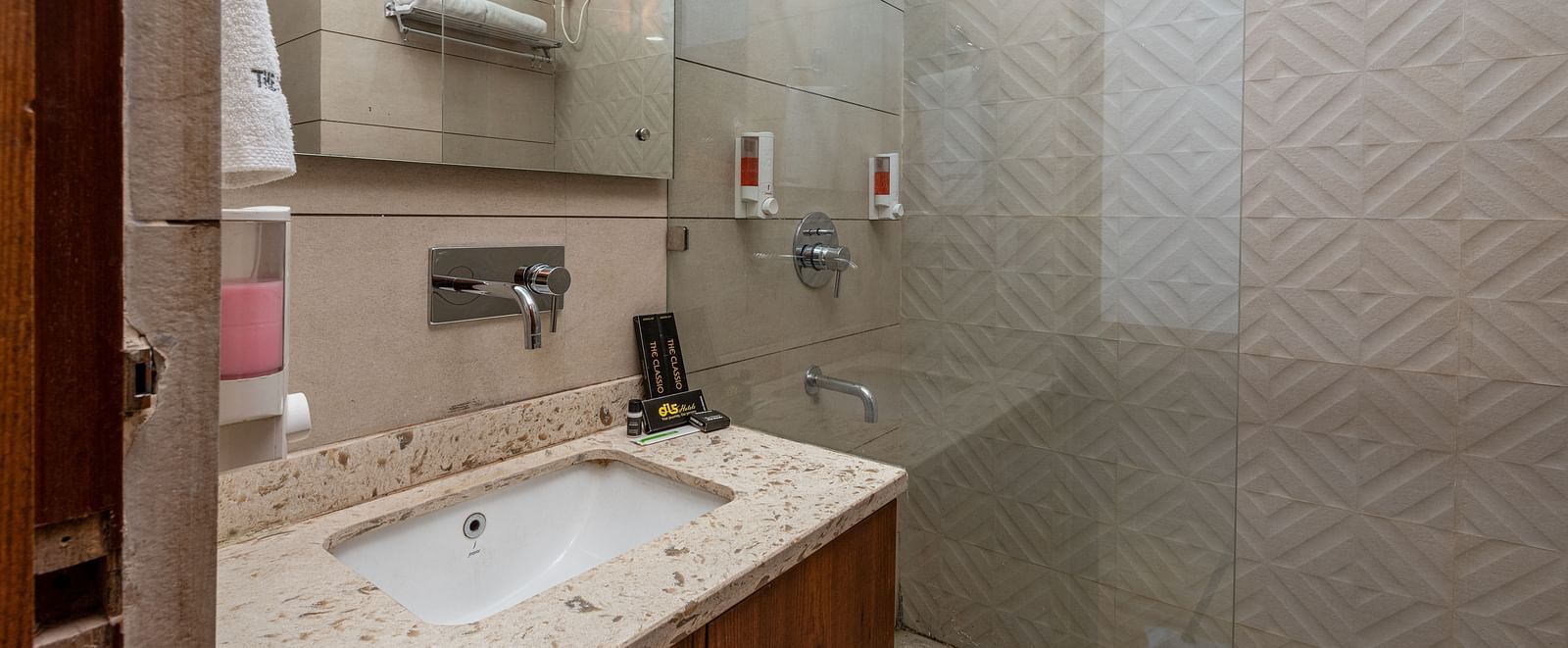 Side view of a bathroom showcasing a mirror above a washbasin with a wooden cabinet beneath and a shower space in the background at DLS The Classio